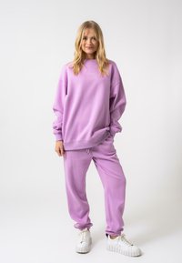 Fialová mikina a odpovídající tepláky vyrobené z měkké tkaniny. Kalhoty mají elastické manžety a obě mají volný střih. Bílé tenisky dokončují outfit.