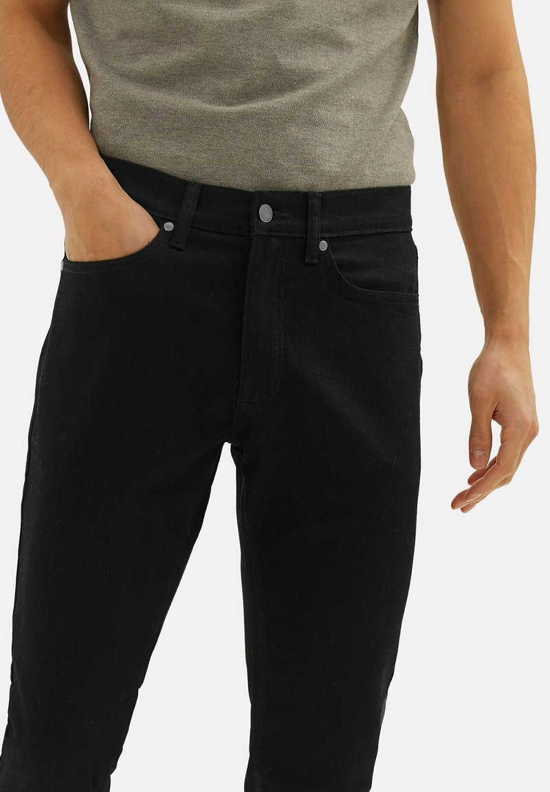 Homme portant un jean noir et une chemise grise, main gauche dans la poche, bras droit détendu le long du corps, devant un fond blanc uni.
