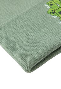 Grøn strikket tæppe med et pixeliseret grafik af en karakter i en kontrasterende farve. Tekstureret stof og foldet kant synlig.