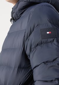 Navy puffer jacket with a quilted design, featuring a high collar and a black patch with logo on the sleeve. Smooth texture and durable fabric.