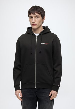Ung mann iført en svart hettegenser med glidelås og "tommy jeans" logo, står foran en ensfarget hvit bakgrunn.