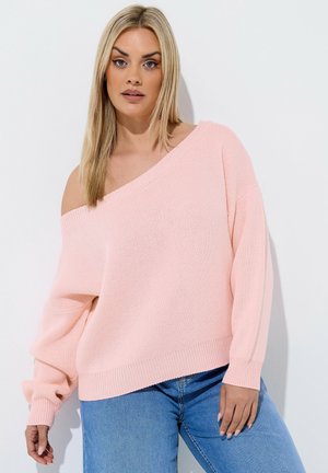 Lyshåret kvinde iført løs lyserød off-shoulder sweater og blå jeans, poserer mod en hvid baggrund.