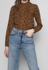 Braunes Hemd mit Knöpfen und schwarzen abstrakten Flecken, langen Ärmeln und einem Stehkragen, kombiniert mit hellblauen hochgeschnittenen Jeans.