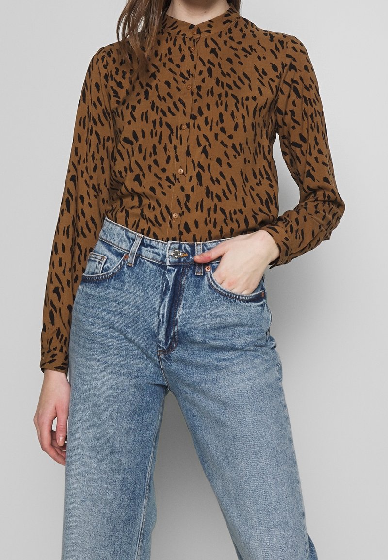 Braunes Hemd mit Knöpfen und schwarzen abstrakten Flecken, langen Ärmeln und einem Stehkragen, kombiniert mit hellblauen hochgeschnittenen Jeans.