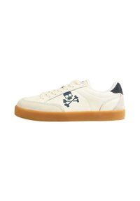 Baskets en suede blanches avec une semelle en caoutchouc marron, arborant un logo de tête de mort et os croisés bleu marine sur le côté, et un accent de talon noir.