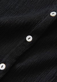 Zwarte gestructureerde stof met een golvend patroon, met verschillende zichtbare witte knopen. De stof lijkt lichtgewicht en iets gekreukt.