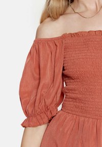 Abito color ruggine senza spalle con maniche a sbuffo, caratterizzato da un corpetto smock e dettagli a volant, realizzato in un tessuto leggero e testurizzato.