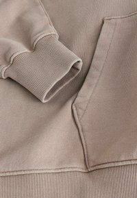 Sudadera beige con puños acanalados y bolsillo. La tela parece suave, con una textura lisa y un diseño minimalista.