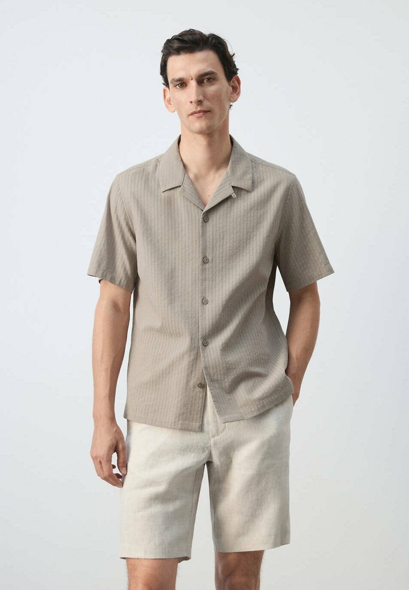 Homme portant une chemise beige à manches courtes boutonnée et un short de couleur claire, debout devant un fond uni.