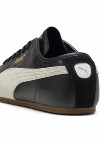 Baskets en cuir noir avec une bande en daim blanc, talon noir texturé et logo Puma doré sur le côté et le talon. Semelle extérieure en caoutchouc.