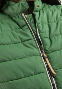 Doudoune matelassée verte avec un col montant, fermeture éclair frontale zippée avec un zip noir contrastant, et détail de tirette en cuir.