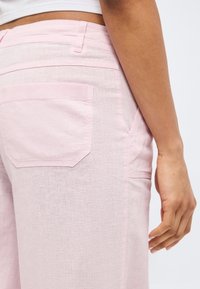 Κοντινό πλάνο της μέσης και του χεριού ενός ατόμου που φοράει ελαφρύ ροζ λινό παντελόνι με πίσω και πλαϊνές τσέπες και λευκό κοντό τοπ.