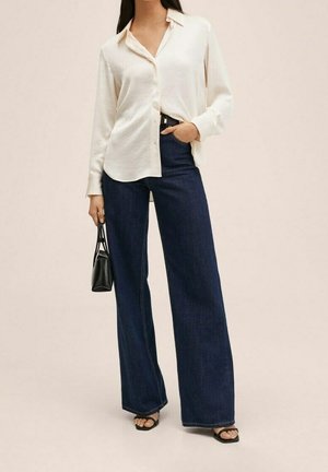 Femme portant une chemise blanche ample boutonnée, un jean large foncé, des sandales noires à talons, et tenant un petit sac à main noir avec une main dans la poche.