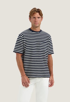 Uomo con capelli castano chiaro indossa una maglietta a maniche corte a righe blu navy e bianca e pantaloni bianchi, in piedi su uno sfondo beige.