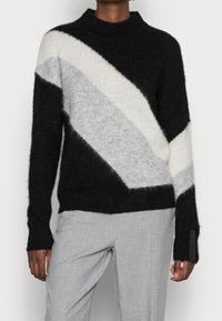 Pull tricoté noir, blanc et gris clair avec un motif chevron incliné, col montant et poignets côtelés. Tissu doux et texturé.