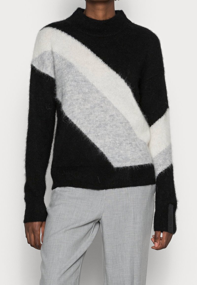 Pull tricoté noir, blanc et gris clair avec un motif chevron incliné, col montant et poignets côtelés. Tissu doux et texturé.