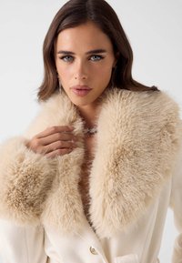 Cappotto color crema con colletto di pelliccia ecologica beige soffice e vaporoso. Presenta bottoni dorati, tessuto strutturato e dettagli morbidi e voluminosi.
