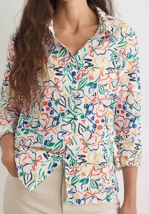 Femme portant une chemise blanche à manches longues avec des motifs floraux abstraits colorés en rouge, bleu, vert, jaune et orange, rentrée dans un pantalon beige.