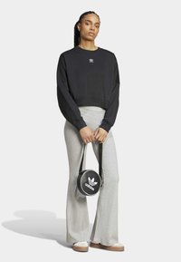 Černý zkrácený svetr s dlouhými rukávy, doplněný světle šedými zvonovými kalhotami. Drží kulatou černou tašku s bílým logem Adidas.