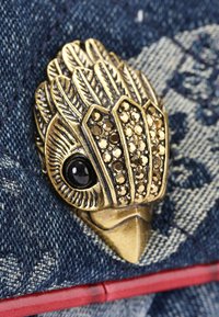 Spilla in metallo dorato a forma di uccello, con dettagli in piuma e accentuazioni di gemme nere, montata su uno sfondo di denim blu texturizzato.
