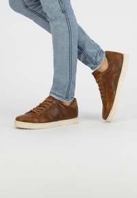 Bruna lädersneakers med beige gummisula, med snörning, texturerade detaljer och en minimalistisk design. Matchas med ljusblå jeans.