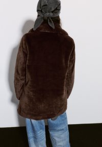 Veste marron duveteuse avec une texture douce, dotée d'un col haut et d'une coupe décontractée. Portée avec un jean large déchiré de couleur bleu clair et un foulard gris.