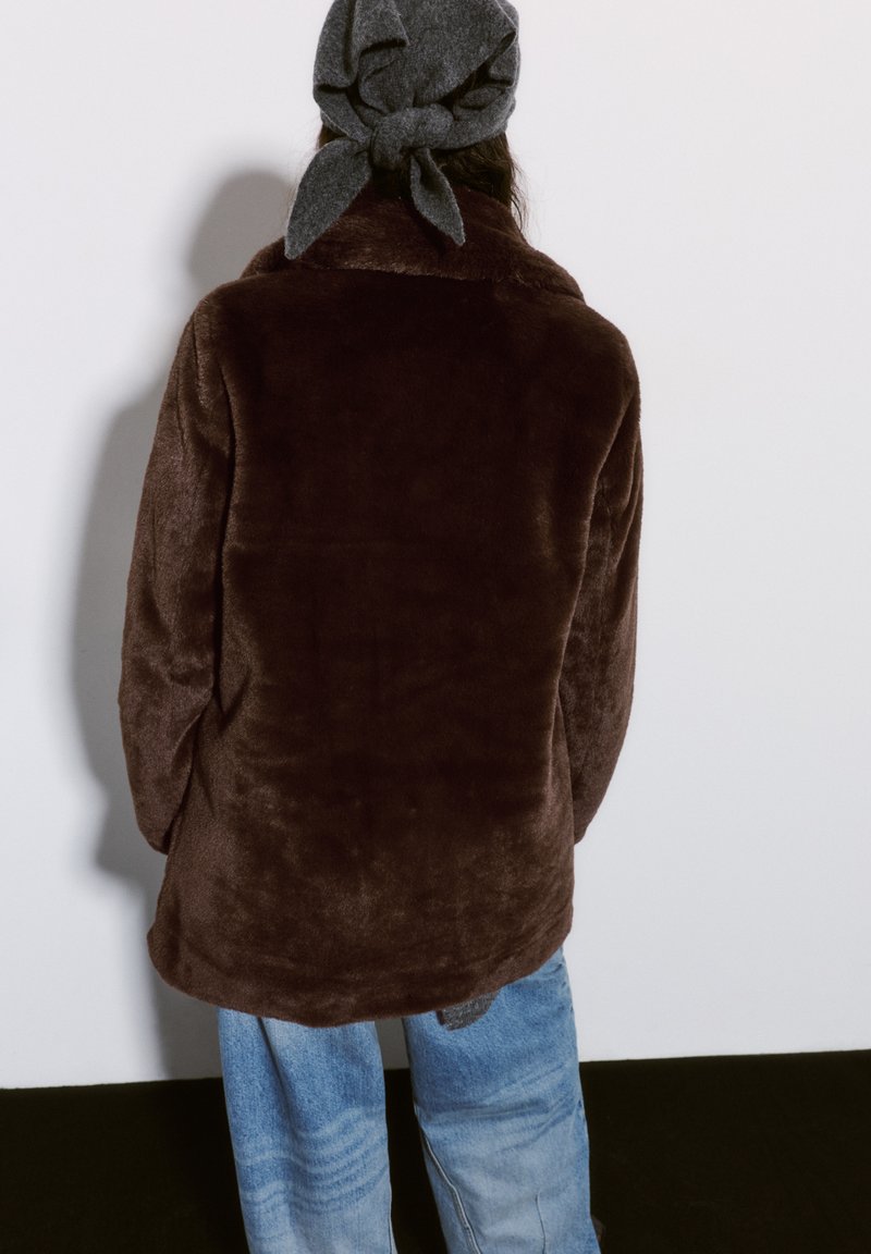 Braune, flauschige Jacke mit einer weichen Textur, die einen hohen Kragen und einen lässigen Schnitt hat. Getragen mit hellblauen, ausgebleichten Weitbeinen-Jeans und einem grauen Kopftuch.