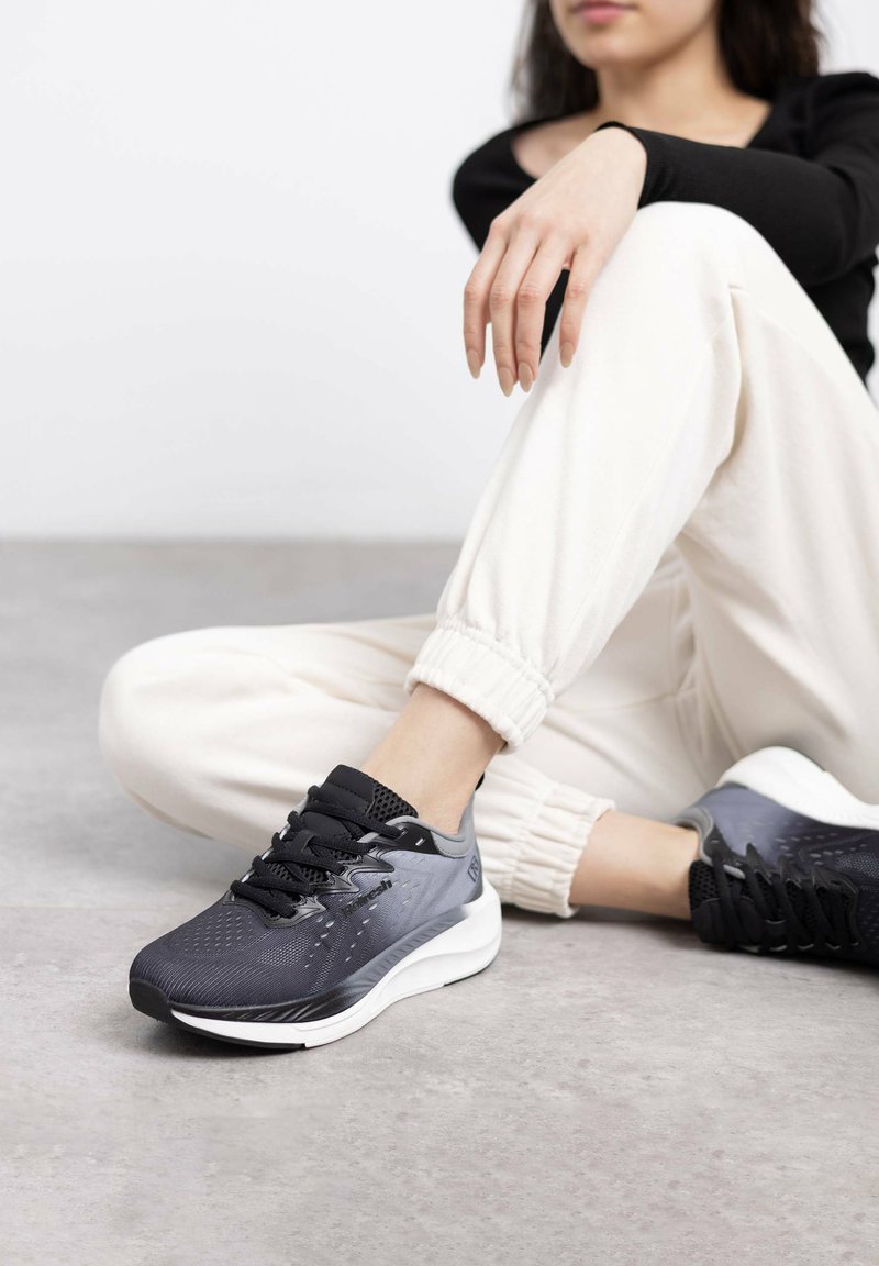 Zapatillas de correr en negro y gris con parte superior de malla y patrones texturizados, cordones y una suela blanca acolchada. Se llevan con pantalones de chándal color crema.