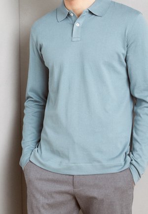 Long-sleeve polo in light blue cotton, featuring a collared neck and two-button placket. Paired with gray trousers. Smooth texture.