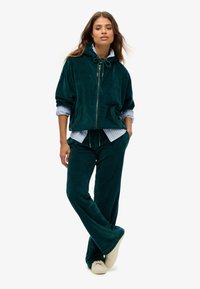 Chándal de terciopelo en color aguamarina con chaqueta con capucha y cremallera, y pantalones de pierna ancha, que cuenta con un cordón y bolsillos laterales, superpuesto a una camiseta de rayas azules.