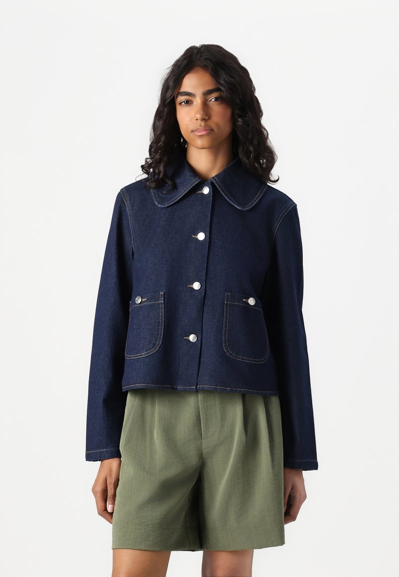 Veste en denim foncé cropped avec un grand col, fermeture devant à boutons, et deux poches plaquées. Des surpiqûres contrastées ajoutent des détails.
