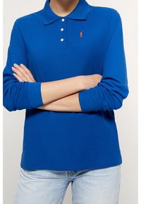 Koszulka polo z długim rękawem w intensywnym niebieskim kolorze, z trzema guzikami przy dekolcie oraz małym pomarańczowym logo na piersi. Miękki materiał o gładkiej teksturze.
