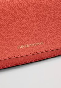 Portafoglio in pelle rossa con texture e logo "Emporio Armani" in rilievo dorato sulla parte inferiore. Presenta cuciture lungo i bordi.