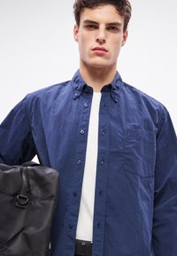 Cămașă bleumarin cu nasturi, cu finisaj texturat, având un buzunar la piept, asortată cu o geantă neagră din piele. Design simplu și linii curate.