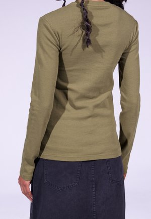 Person med fletninger iført en olivengrøn langærmet skjorte og en mørk denimnederdel, set bagfra.