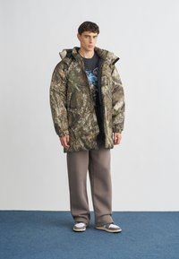 Giacca a vento mimetica, toni terrosi e motivi di corteccia d'albero, abbinata a pantaloni grigi larghi e sneaker bianche con dettagli grigi.