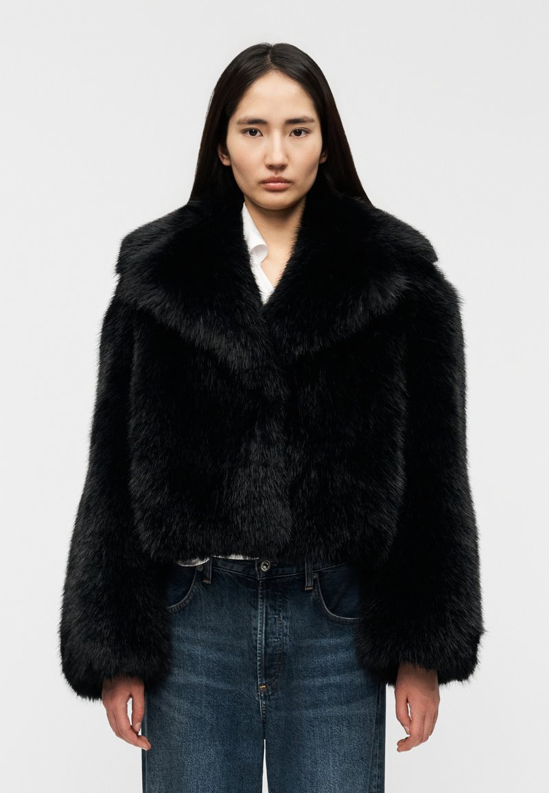 Μαύρο κοντό μπουφάν από συνθετική γούνα με φαρδι ά γιακά, μακριά μανίκια και απαλή υφή, σε συνδυασμό με μπλε τζιν παντελόνι.