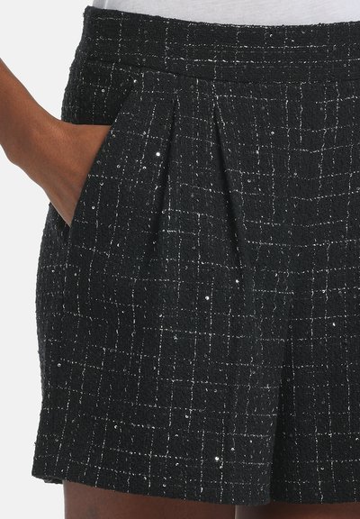 Shorts en tweed noir avec une surface texturée, présentant un motif subtil de fils métalliques et des poches latérales. Un détail plissé à l'avant rehausse la forme.