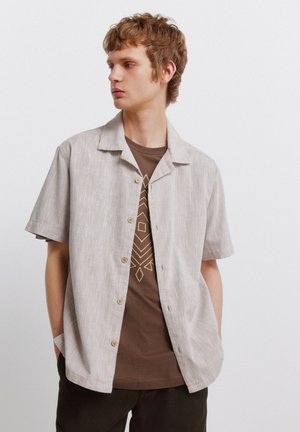 Junger Mann mit lockigem Haar, der ein beiges kurzärmeliges Hemd über einem braunen T-Shirt mit geometrischem Muster trägt.