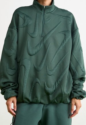 Personne portant un pull matelassé vert foncé avec de grands motifs brodés en forme de virgule et un pantalon vert assorti avec un petit logo près de la poche.