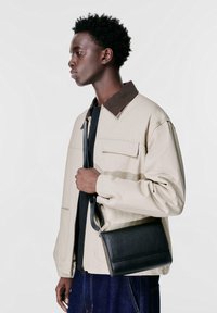 Giacca beige con zip e colletto marrone, indossata sopra una maglietta nera. Borsa a tracolla in pelle nera di forma rettangolare, portata sul lato sinistro.