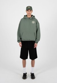 Mees päevas rohelist kapuutsi ja sobivat mütsi, mustad lühikesed püksid, valged sokid ja mustad jalanõud, seisab ühevärvilise heleda tausta ees.