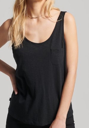 Kobieta ubrana w czarny bezrękawnik z dekoltem typu scoop i małą kieszonką na piersi, stojąca na tle jednolitego szarego tła.