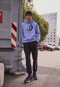 Svetle modrá mikina s kapucí a logem, černé tepláky s brandingem a černé tenisky. Pozadí zahrnuje šedou nádobu a městskou scenérii.