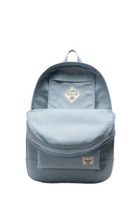 Un sac à dos bleu clair en tissu, de forme arrondie, avec un rabat à l'avant, une poignée sur le dessus, et un petit logo sur la poche avant.