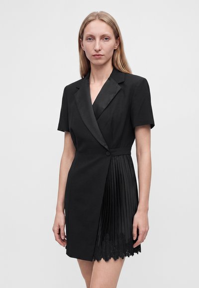 Femme aux longs cheveux blonds porte une robe noire à manches courtes avec un revers en satin et une jupe plissée ornée de dentelle sur un fond uni.