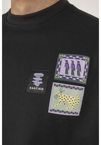 Felpa nera con tre toppe di stoffa colorate: un logo a forma di serpente, uccelli blu e un gatto giallo con punti, il tutto su uno sfondo texturizzato.