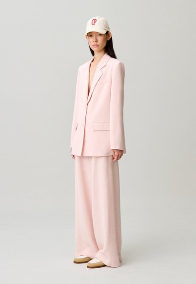 Femme debout portant un blazer oversize rose clair et un pantalon large, une casquette beige avec des initiales rouges, et des mocassins beige.
