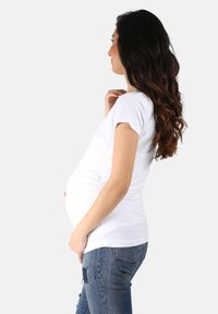 Weißes Schwangerschafts-T-Shirt mit kurzen Ärmeln, gerafften Seiten für Stretch, kombiniert mit hellblauen, verwaschenen Jeans. Glattes Material und lässiges Design.