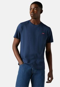 Giovane uomo che indossa una maglietta Levi's blu navy e jeans blu, con una mano in tasca, che guarda di lato su uno sfondo uniforme.